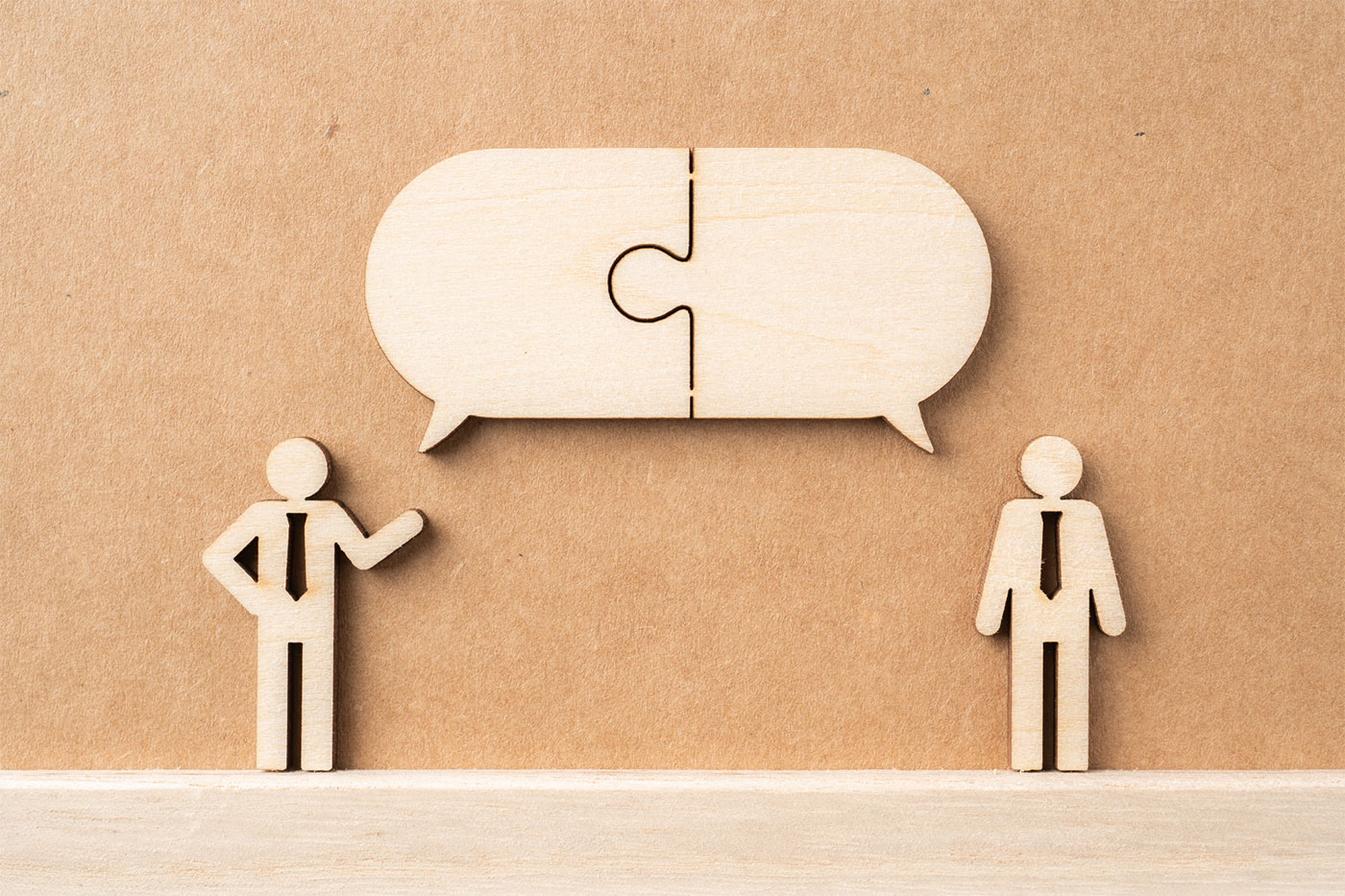 Two wooden figurines depicting people standing, with one figure raising an arm, positioned alongside a speech bubble crafted from puzzle pieces.