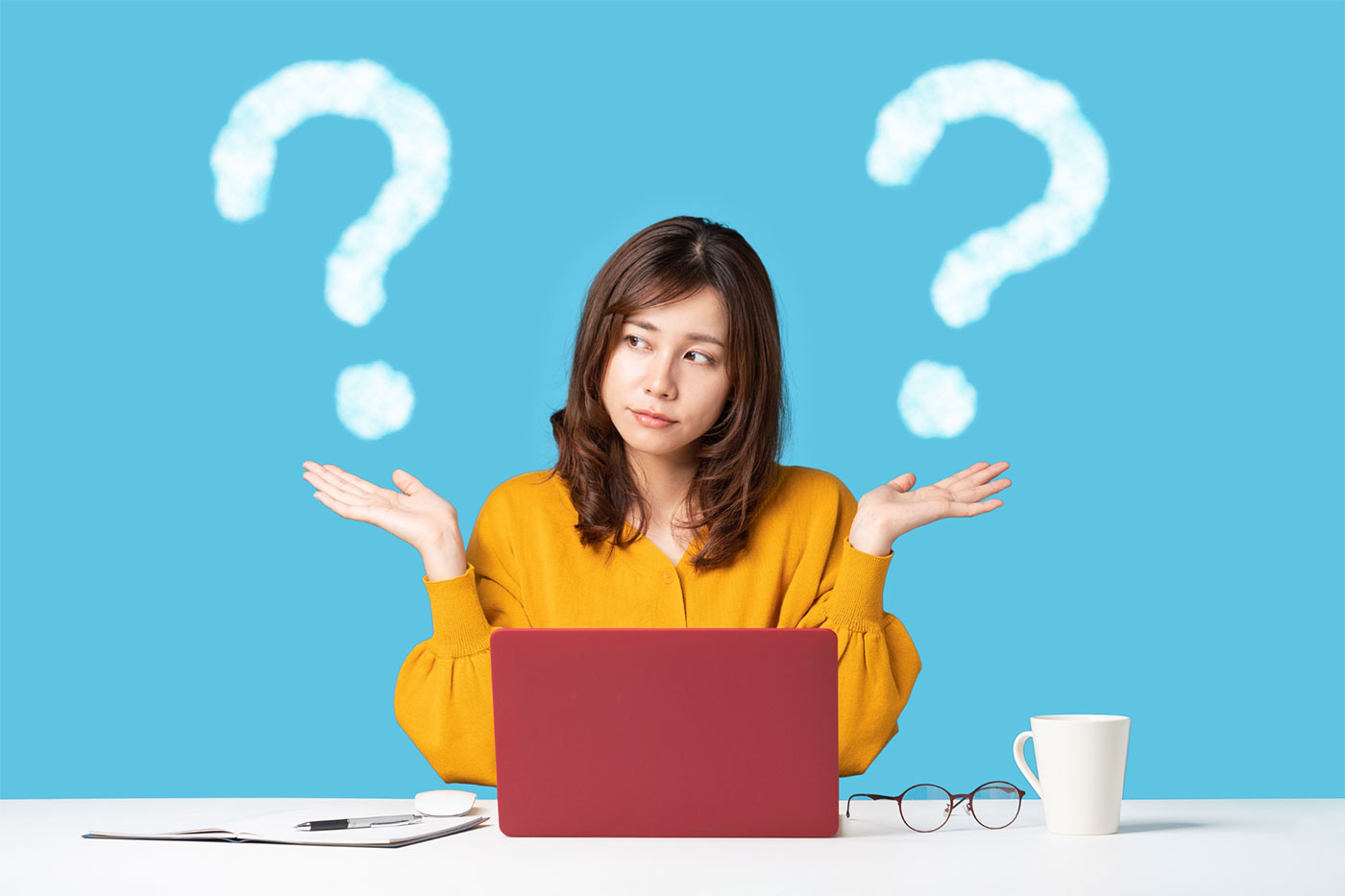 A person seated at a desk with a laptop and a cup, raising their hands in a shrugging gesture, with two question marks above them.