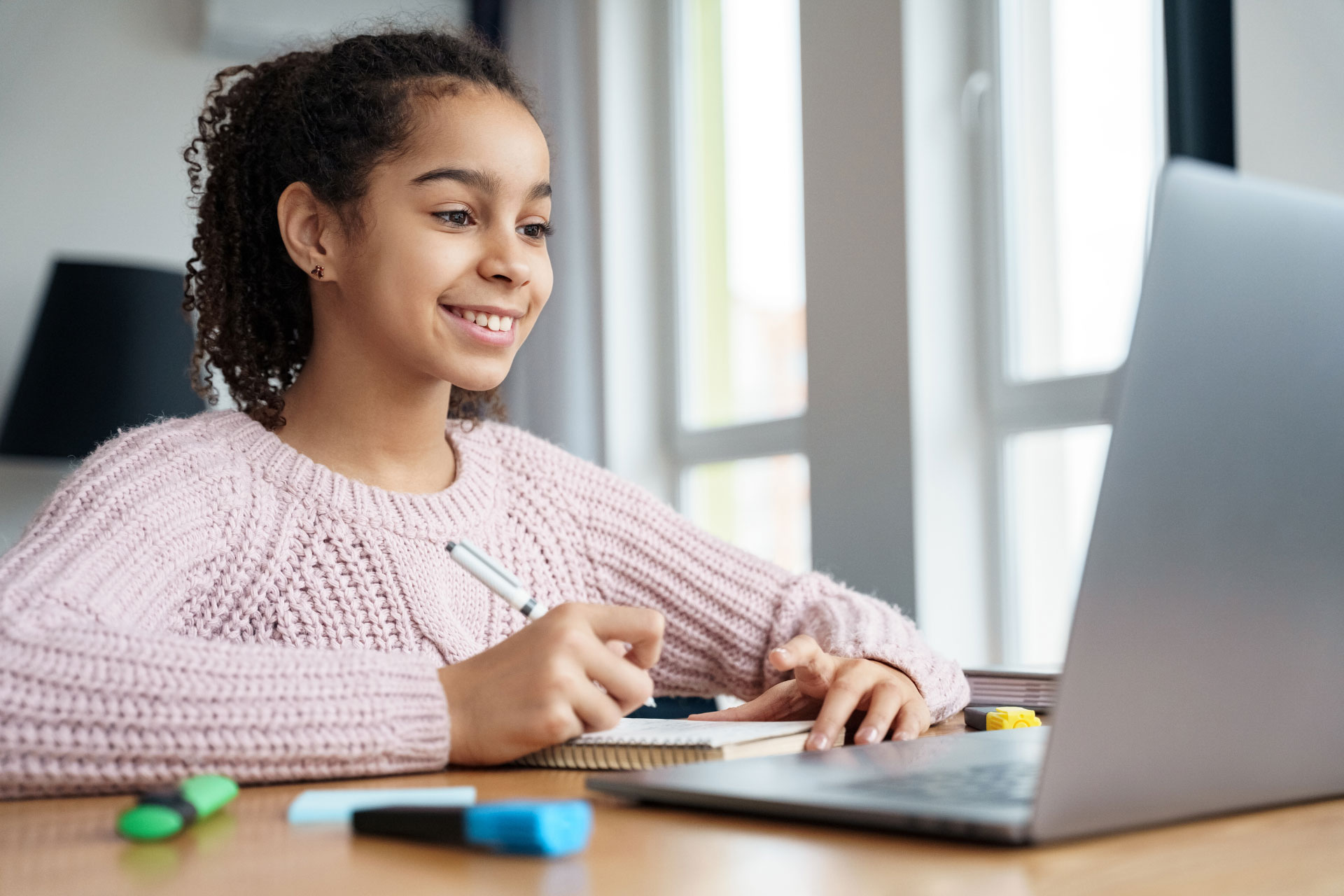 A young student in a pink sweater taking an online class, pen and notepad in hand