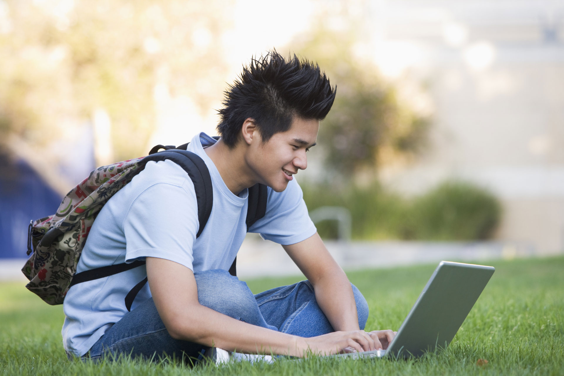 A young man wearing a backpack taking an online class outdoors, sitting on the grass
