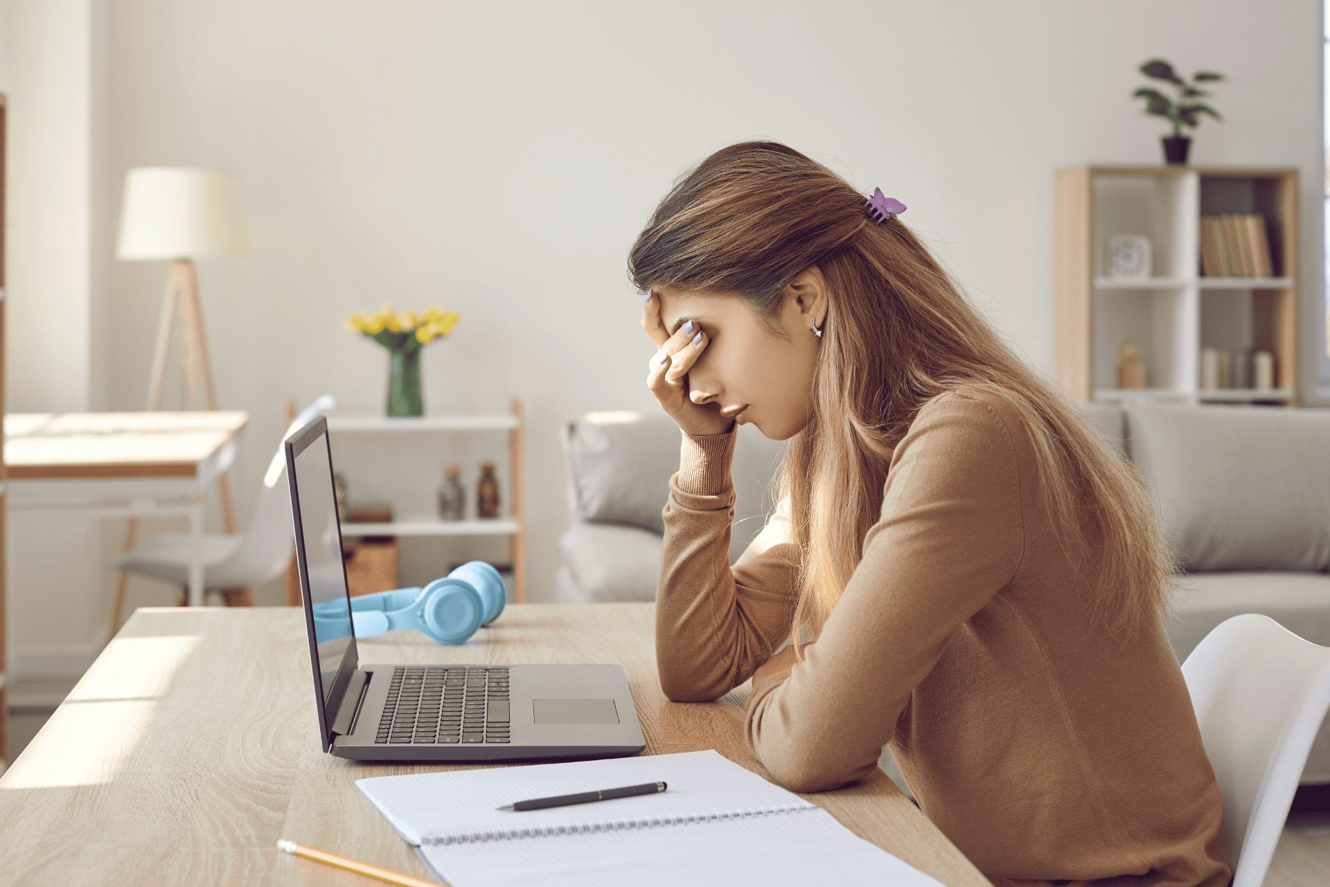 A young, visibly stressed woman sits at a desk with an open laptop and notebook, rubbing her eyes.