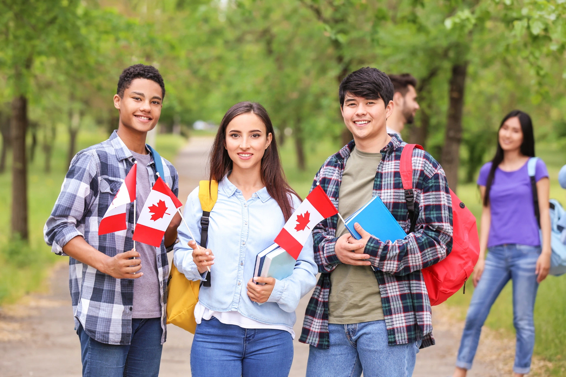 Three international students stand in a park holding books and Canadian flags.