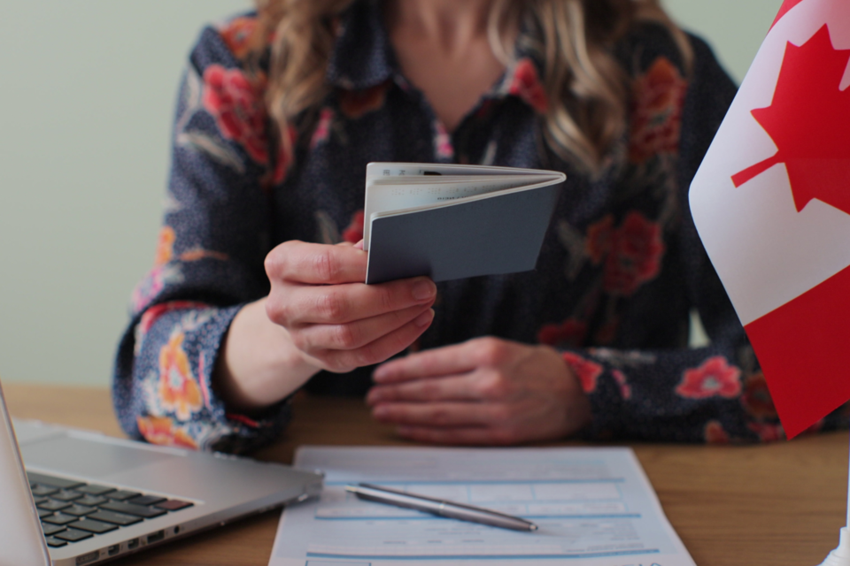 A woman sitting at a desk with a laptop, paper, pen and Canadian flag, holding a passport.