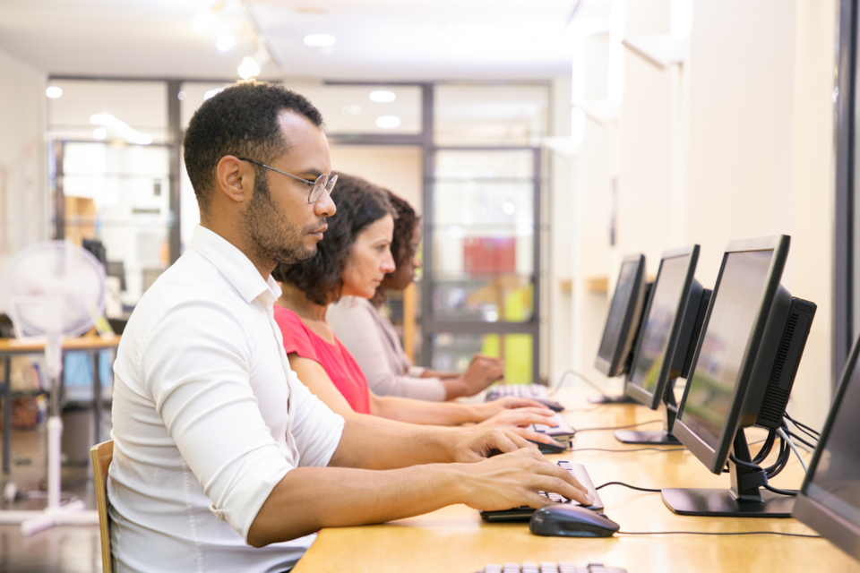Three focused adults are seated at computers taking a CELPIP exam.