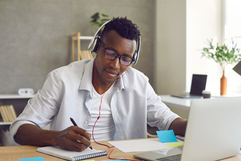 A young man with headphones sits in front of a laptop surrounded by sticky notes.